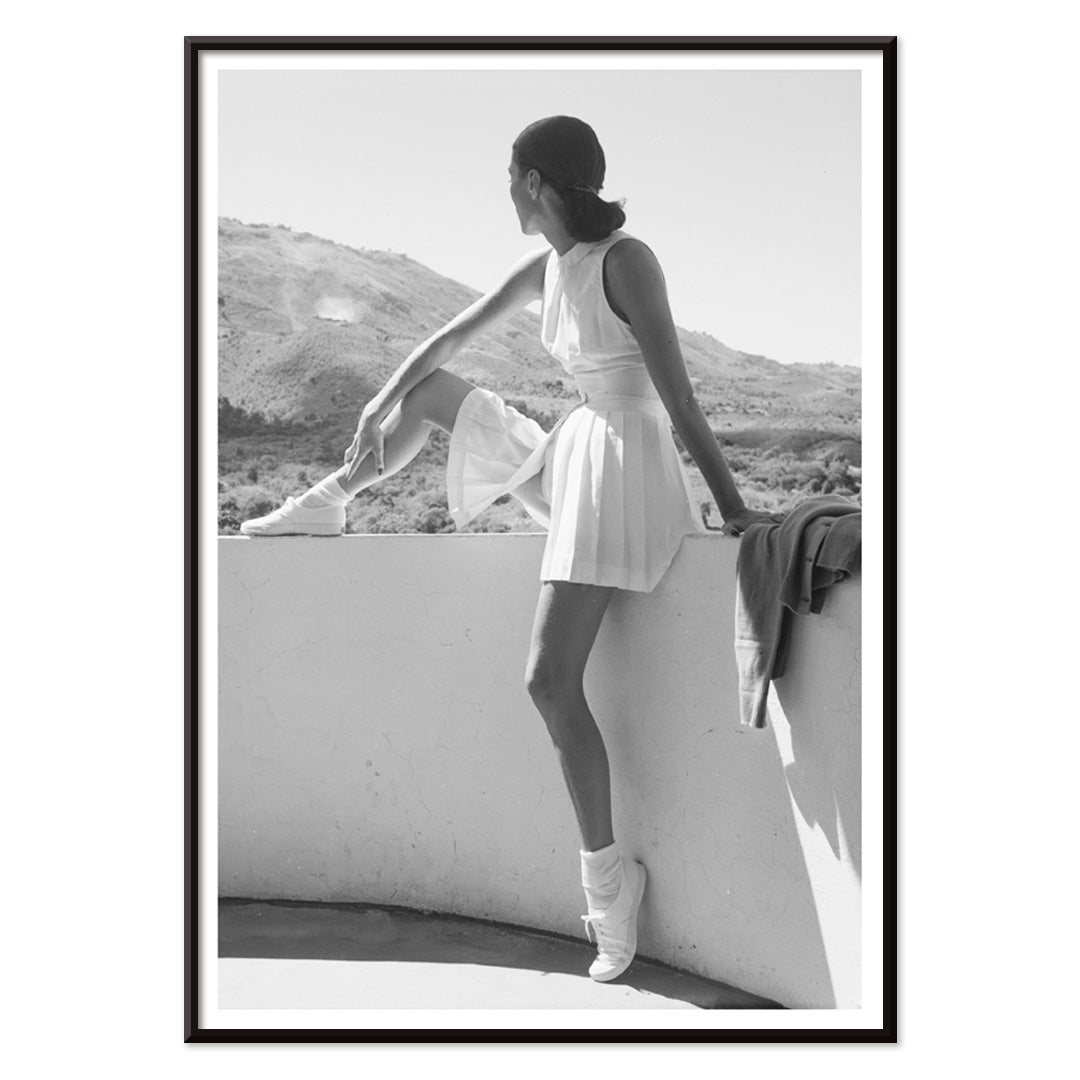 Fashion model in tennis outfit seated on wall looking at mountains by Toni Frissell poster artwork, with black aluminium frame on white background